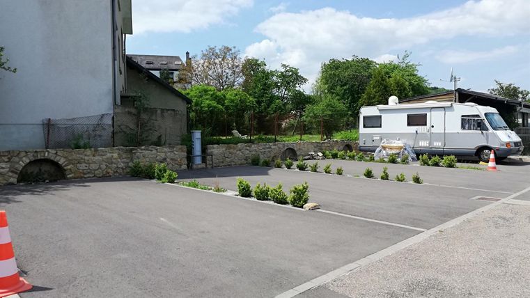 Een parkeerplaats met een paar kleine groenvoorzieningen en een camper. Op de achtergrond zijn bomen en een gebouw zichtbaar.