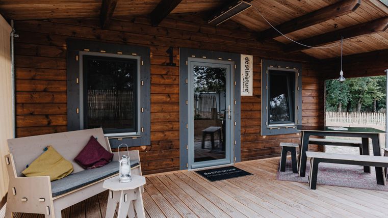 Eine gemütliche Veranda eines Holzhauses mit einer Sitzgelegenheit und einem Tisch. Die Umgebung wirkt ruhig und einladend.
