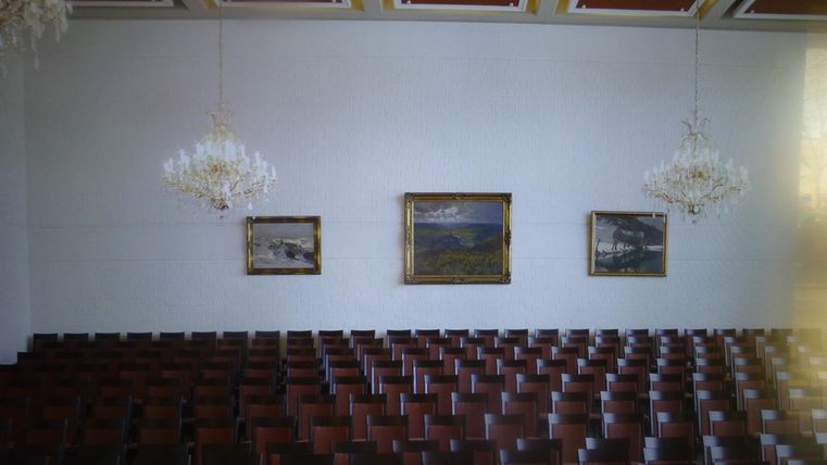 Ein großer Raum mit vielen Stühlen in Reihen. An den Wänden hängen drei Bilder, und es gibt zwei Kronleuchter.