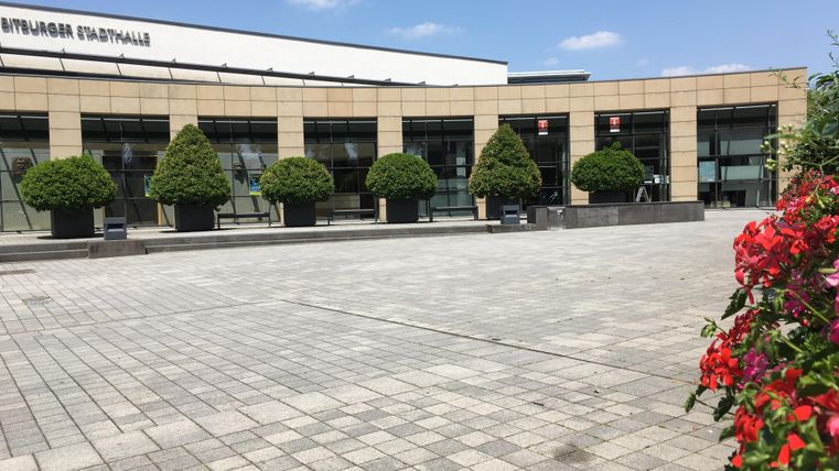 Ein modernes Gebäude mit großen Fenstern und gleichmäßig angeordneten Bäumen. Im Vordergrund blühen farbige Pflanzen auf einem gepflasterten Platz.
