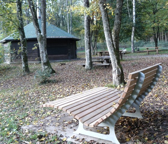 Holzliege und Picknicktische in einem herbstlichen Wald mit einer H&uuml;tte im Hintergrund. Laub bedeckt den Boden., &copy; Felsenland S&uuml;deifel Tourismus GmbH, Christian Calonec-Rauchfuss