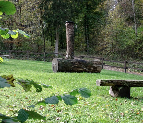 Holzskulptur und Bank auf einer grünen Wiese, umgeben von Wald. Ein Wanderweg führt durch die herbstliche Landschaft., © Tourist-Info Bitburger Land