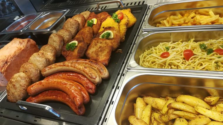 Een selectie van verschillende vleesgerechten, waaronder worstjes en burgers, wordt in een vitrine gepresenteerd. Naast de vleessoorten zijn er ook pasta en aardappelpartjes.