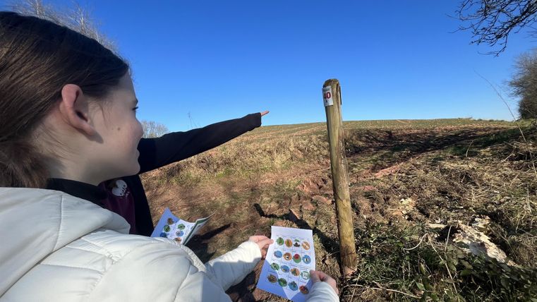 Zwei Personen stehen auf einem Feld und zeigen auf ein Schild. Sie halten Karten mit verschiedenen Symbolen in der Hand.
