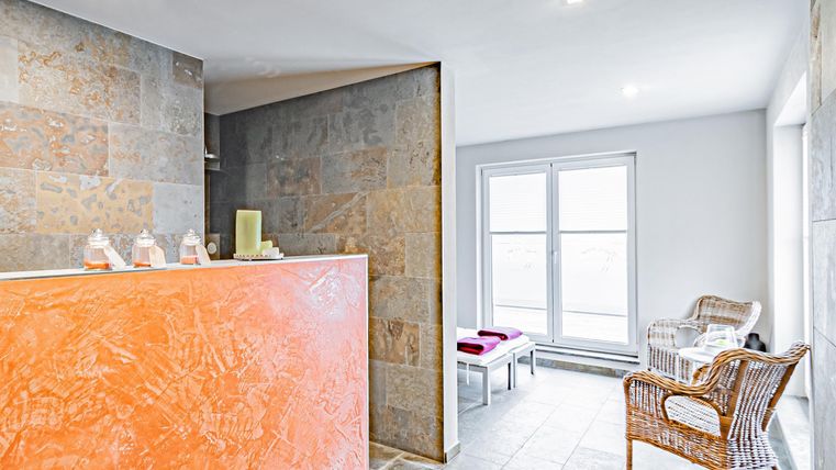 Ein moderner Wellnessbereich mit einem orangefarbenen Empfangstresen. Es gibt gemütliche Rattanstühle und große Fenster, die viel Tageslicht bieten.