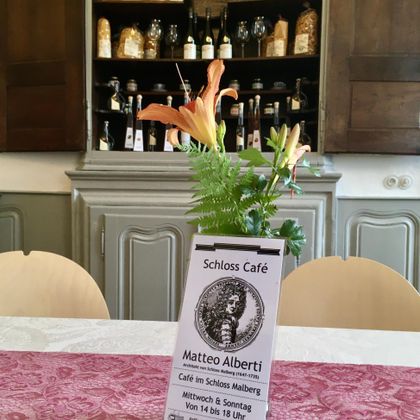 Un intérieur de café accueillant avec une décoration de table florale et une ardoise du menu. En arrière-plan, des étagères avec différentes bouteilles et verres sont visibles.