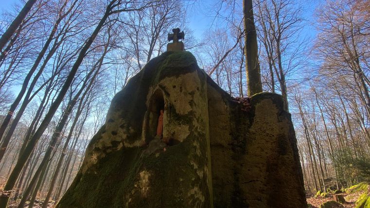 Ein beeindruckender Felsen mit einer kleinen Öffnung und einem Kreuz darauf, umgeben von Bäumen. Der Himmel ist klar und blau, was eine ruhige Atmosphäre schafft.