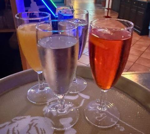 Vier glazen met verschillende dranken op een dienblad. Op de achtergrond zijn diffuse lichten en een stijlvolle inrichting zichtbaar.