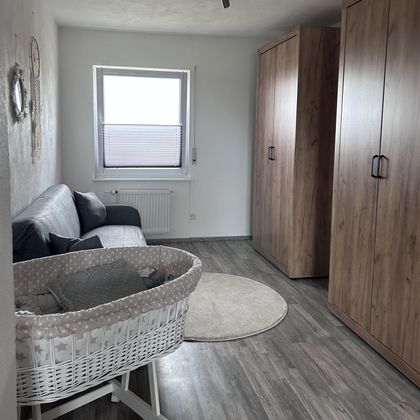 Een heldere kamer met een bank, een tapijt en een babybedje. De kamer heeft houten kastdeuren en een raam dat natuurlijk licht binnenlaat.