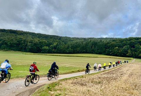 Vier Personen stehen vor einem Café und posieren mit einem E-Bike. Sie lächeln und tragen sportliche Kleidung.