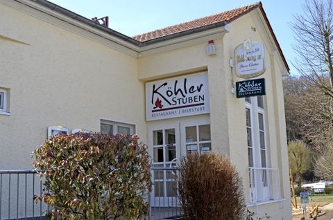 Ein Restaurant mit dem Namen „Köhler Stuben“ ist an einer hellen Wand zu sehen. Vor der Eingangstür stehen einige Pflanzen.