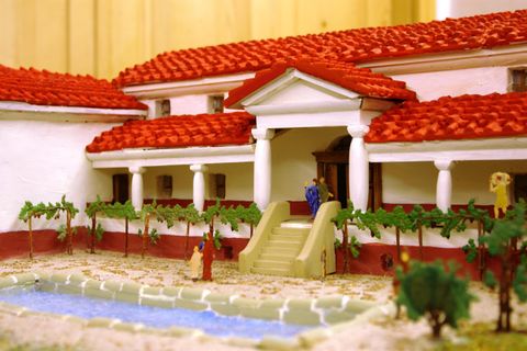 Ein detailliertes Modell eines römischen Hauses mit rotem Ziegeldach. Im Vordergrund sind ein kleiner Teich und Weinreben zu sehen.