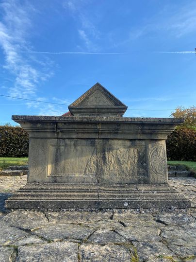 Ein steinernes Grabmal mit einer pyramidenförmigen Spitze steht unter einem blauen Himmel. Der umliegende Boden ist aus großen Steinen und zeigt grüne Vegetation im Hintergrund.