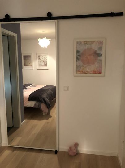 Ein moderner Raum mit einer Schiebetür, die in ein Schlafzimmer führt. An der Wand hängen Bilder und es gibt einen sanften Lichtschein von einer Deckenlampe.