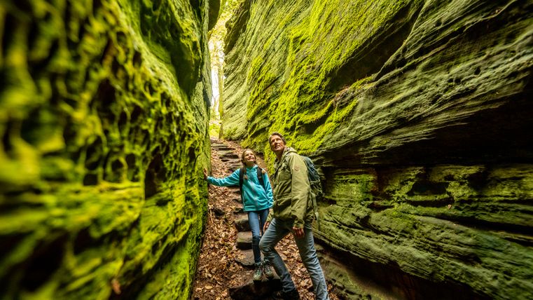 Zwei Personen wandern durch eine enge, moosbedeckte Schlucht im Wald.