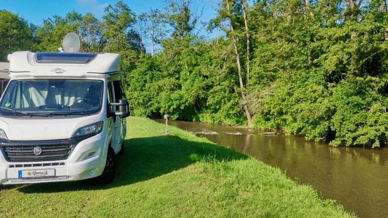 Ein Wohnmobil steht an einem ruhigen Flussufer, umgeben von üppigem Grün. Der Himmel ist klar und blau, ideal für einen entspannten Tag in der Natur.