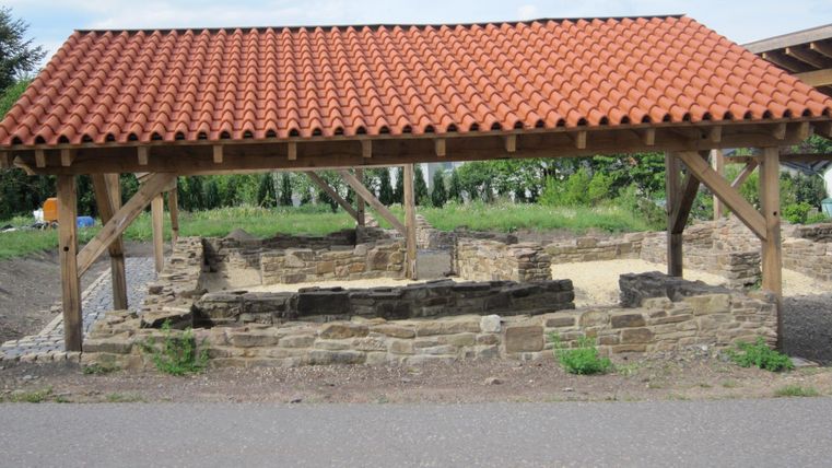 Eine Überdachung aus Holz schützt die Ruinen eines alten Gebäudes. Im Hintergrund sind grüne Wiesen und ein blauer Himmel sichtbar.