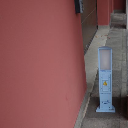 A blue pole stands against a wall, next to a walkway. The pole features a warning symbol and a light strip on top.