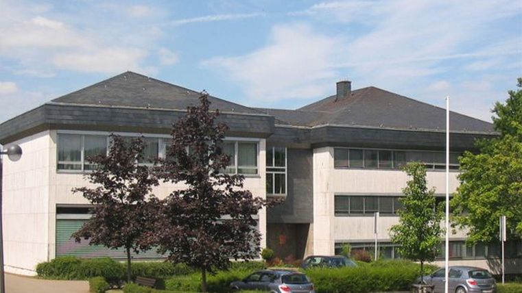 Een modern gebouw met een hellend dak en grote ramen. Omringd door bomen en enkele geparkeerde auto's.