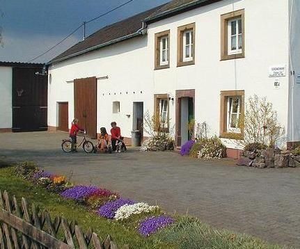 Ein typisches Landhaus mit einer hellen Fassade und einem gepflasterten Hof. Kinder spielen im Freien und Blumen schmücken den Vorgarten.