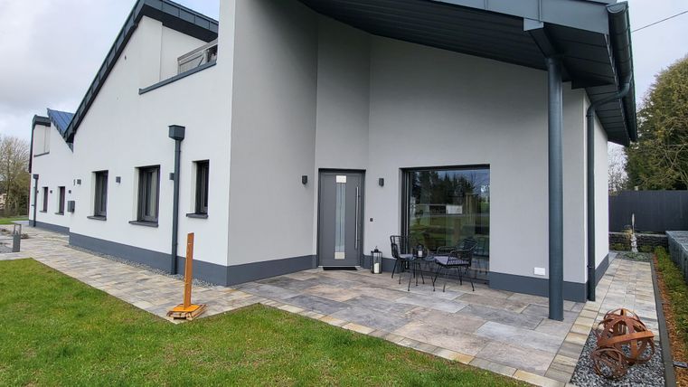 Une maison moderne avec une façade gris clair et un toit plat. Devant la maison, il y a un espace extérieur pavé avec une table et des chaises.