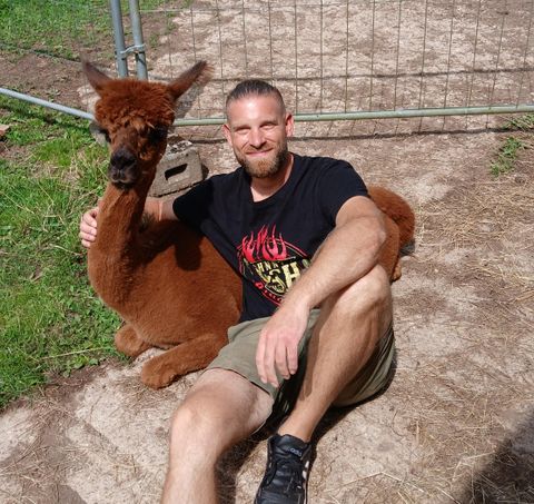 Un homme est assis à côté d'un alpaga sur le sol. Les deux semblent détendus et heureux dans un environnement verdoyant.
