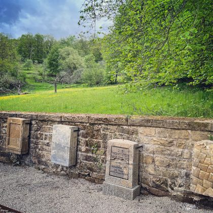 Eine Steinmauer mit mehreren Gedenktafeln. Im Hintergrund sind grüne Wiesen und Bäume zu sehen.