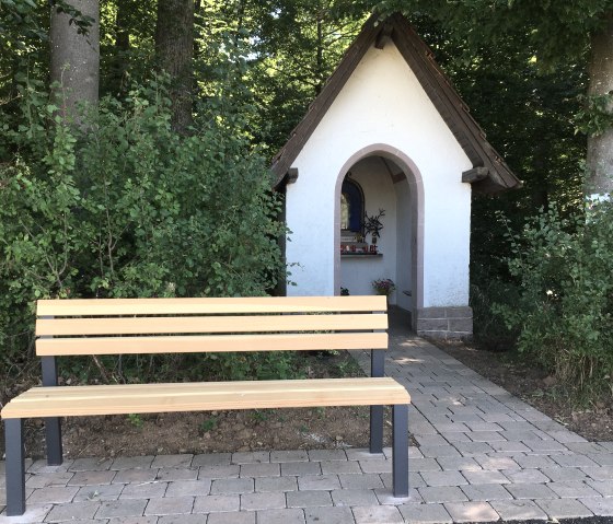 A small white chapel with a brown roof stands at the edge of the forest. In front of it is a wooden bench on a paved path., &copy; TI Bitburger Land