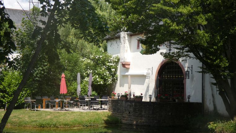 Un restaurant confortable avec une terrasse en plein air, entouré de verdure. Des parasols rouges sont prêts, tandis que des arbres à l'arrière-plan offrent de l'ombre.