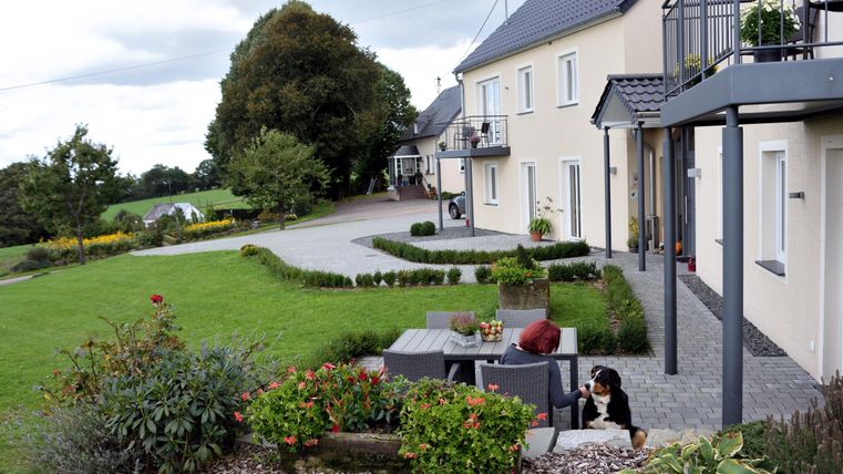 Eine ruhige Wohngegend mit einem modernen Haus und einem gepflegten Garten. Im Vordergrund sind ein Sitzbereich und ein Hund zu sehen.