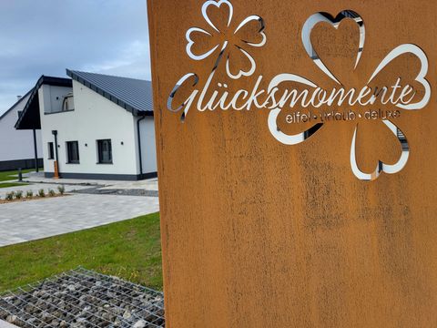 Un complexe moderne avec une enseigne d'entrée rustique affichant les mots "Moments de bonheur". Les environs sont bien entretenus avec de l'herbe verte et des surfaces en pierre.