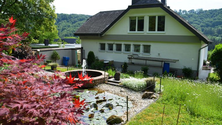 A beautiful house with a well-maintained garden and a pond. In the background, gentle hills and trees can be seen.