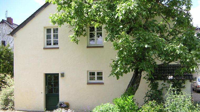 Ein gemütliches Haus mit gelber Fassade und großen Fenstern. Vor dem Haus steht ein großer Baum und blühende Pflanzen.