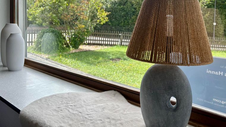 Eine stilvolle Lampe mit einem runden, grauen Sockel und einem schilfgefertigten Lampenschirm steht auf einem Tisch. Draußen ist eine grüne Wiese und ein Zaun zu sehen.
