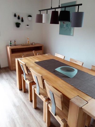 Ein moderner Essbereich mit einem großen Holztisch und Stühlen. Über dem Tisch hängen stilvolle Lampen in verschiedenen Farben.