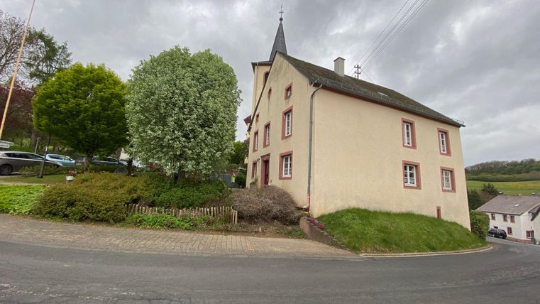Een charmant, tweelaags huis aan een kronkelige straat. Rondom het gebouw staan bomen en een groene weide.