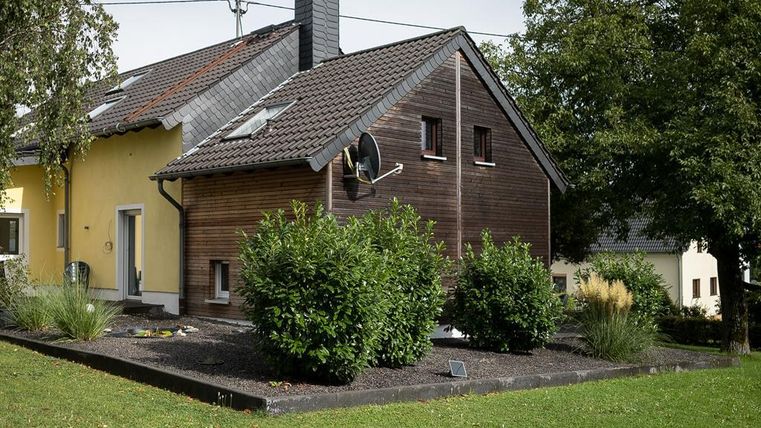 Une maison moderne avec une façade en bois et une façade jaune. Le jardin est aménagé avec des buissons et du gazon.