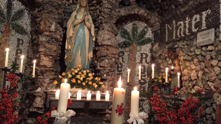 Ein Andachtsort mit einer Statue der Jungfrau Maria, umgeben von Kerzen und Blumen. Die Wände sind mit Steinen verziert und zeigen mosaikartige Verzierungen.