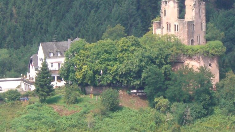 Une ancienne ruine de château, entourée d'arbres, se dresse sur une colline. Au premier plan se trouve un bâtiment d'hôtel.