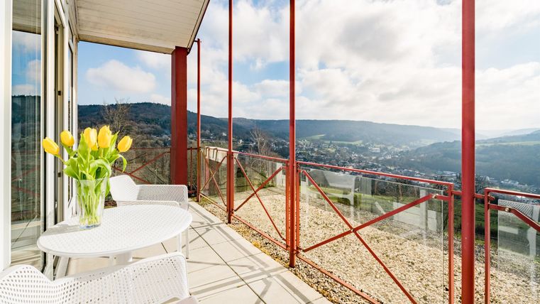 Ein sonniger Balkon mit einem weißen Tisch und Stühlen. Im Hintergrund erstreckt sich eine malerische Aussicht auf grüne Hügel und einen bewölkten Himmel.