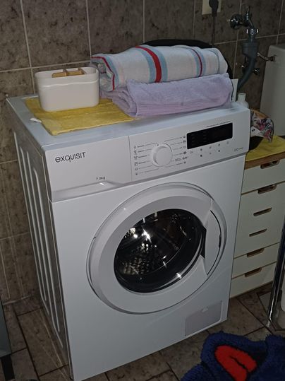 Eine Waschmaschine steht in einem Badezimmer. Handtücher liegen ordentlich darauf und die Wände sind gefliest.