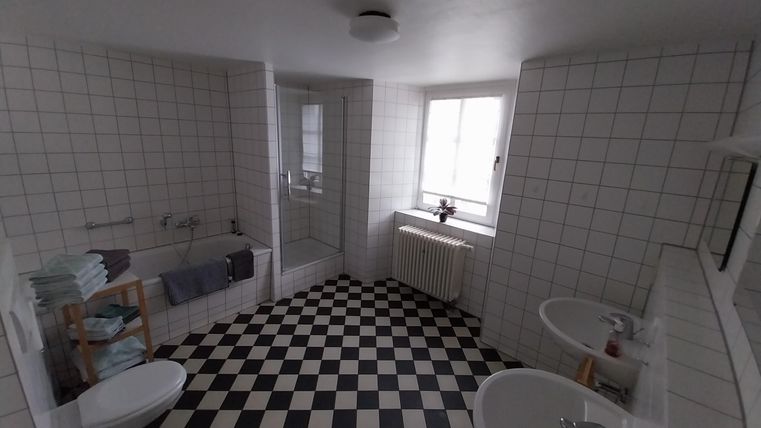 Ein helles Badezimmer mit schwarz-weißen Fliesen. Es enthält eine Badewanne, zwei Waschbecken und ein Fenster für natürliches Licht.