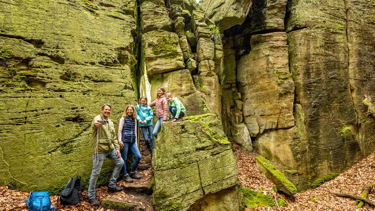 A group of hikers stands in an impressive rocky landscape. The green rocks are covered in moss and surrounded by autumn foliage.