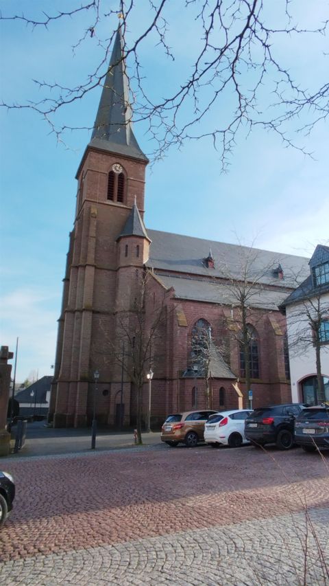 Eine alte Kirche mit einem hohen Turm steht im Vordergrund. Im Hintergrund sind einige Fahrzeuge und eine klare blaue Himmel zu sehen.