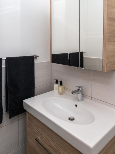 Ein modernes Bad mit einem Waschbecken und einem großen Spiegel. Die Ausstattung umfasst Handtücher und Pflegeprodukte auf der Ablage.