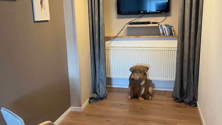 Une pièce confortable avec deux chaises et un pouf. Un téléviseur est accroché au mur, et un chien est assis au milieu de la pièce.