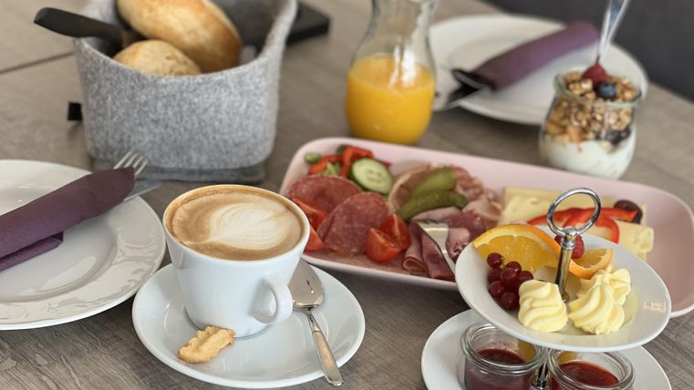 A hearty breakfast with coffee, fresh rolls, cold cuts, and fruit. Next to a glass of orange juice, there is a yogurt with muesli.
