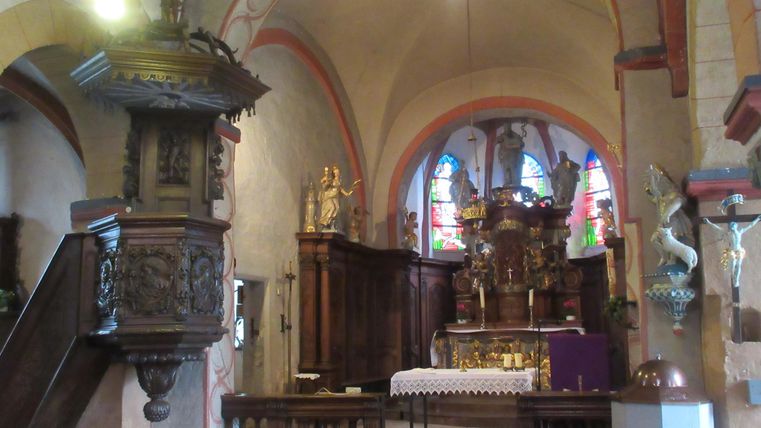 Ein Innenraum einer Kirche mit einem kunstvoll geschnitzten Kanzel und bunten Fenstern. Der Altar ist im Hintergrund sichtbar, umgeben von Holzverkleidungen.