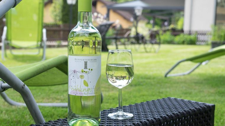 Eine Flasche Weißwein steht neben einem Glas Wein auf einem Tisch im Garten. Im Hintergrund sind Liegestühle und eine grüne Wiese zu sehen.