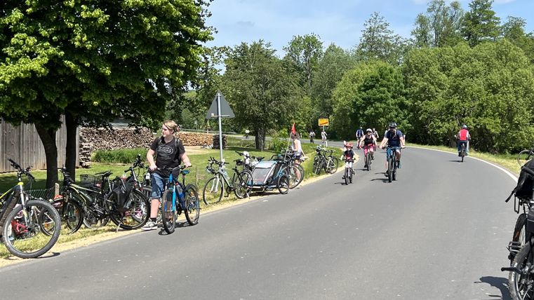 Eine Straße mit Radfahrern und parkierten Fahrrädern. Die Umgebung ist grün und sonnig.